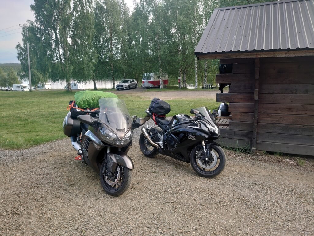 Kaksi moottoripyörää (Kawasaki ja Suzuki) parkissa Rantalan majoitusalueella, taustalla järvi ja leirintäalue.